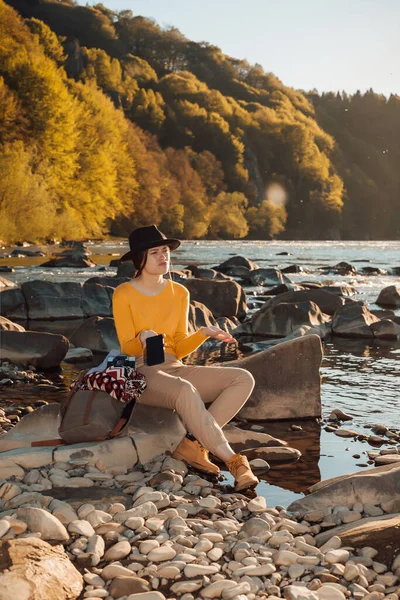 Young woman traveler with cell phone in hands sitting on rock near river nature