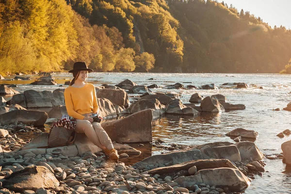 Young woman traveler with cell phone in hands sitting on rock near river nature