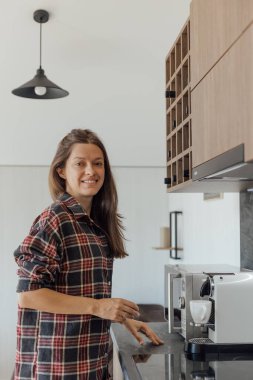 Bir kadın şık mutfağında duruyor, kahveyi hazırlarken gülümsüyor. Ekose bir gömlek giyiyor ve mutfakta parlak aletler ve sıcak ışıklandırma var..