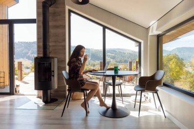 Geniş pencereli parlak bir odada, küçük yuvarlak bir masada dizüstü bilgisayarda çalışan bir kadın. Dağların ve yeşilliklerin manzarası hoşuna gidiyor. Huzurlu bir atmosfer yaratıyor..