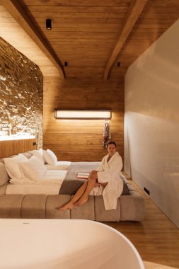 Şık bir otel odasında, bornoz giymiş bir kadın sakin atmosferin tadını çıkarır. Doğal malzemeler ve yumuşak ışıklandırma dinlenmek için sıcak ve davetkar bir alan yaratır..