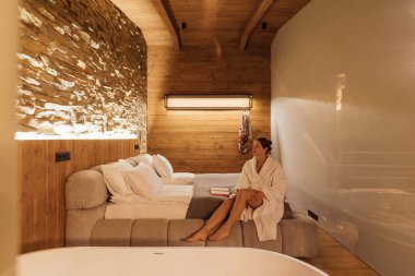 Şık bir otel odasında, bornoz giymiş bir kadın sakin atmosferin tadını çıkarır. Doğal malzemeler ve yumuşak ışıklandırma dinlenmek için sıcak ve davetkar bir alan yaratır..