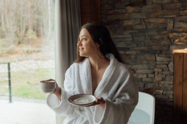 Kabarık beyaz bornozlu bir kadın, güzel doğal bir manzaraya bakarken sıcak bir fincan kahvenin tadını çıkarıyor. Yanında bir tabak tutuyor ve sakin bir sabah atmosferi yaratıyor..