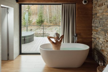 Bir insan şık bir küvette huzurlu bir andan zevk alır, saçlarını havluyla nazikçe kurutur, dingin ahşap ve taş detaylarla çevrili, doğal ışık odayı su basar..