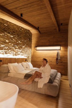 Şık bir otel odasında, bornoz giymiş bir kadın sakin atmosferin tadını çıkarır. Doğal malzemeler ve yumuşak ışıklandırma dinlenmek için sıcak ve davetkar bir alan yaratır..