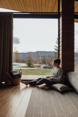 Büyük bir pencerenin yanında dizüstü bilgisayarıyla çalışan bir kadın oturuyor. Sıcak ahşap iç mekan rahat bir atmosfer yaratırken o dışarıdaki doğanın tadını çıkarıyor..