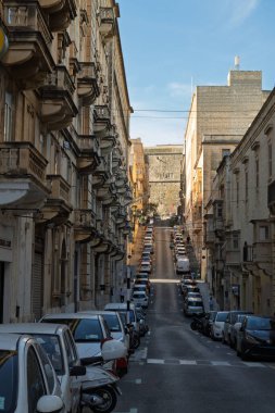 Valletta, Malta 'da dik bir cadde park etmiş arabalar ve yayalarla dolu. Mimari tarihi, balkonları var. İnsanlar şehri keşfetmek için sokakta yürüyorlar..