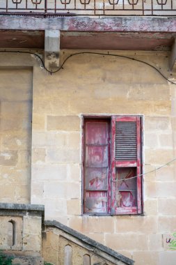 Eski bir bina yaşını gösterir. Kırmızı pencere pervazı soyulmuş ve cam kırılmış. Bina taştan yapılmış ve merdivenler pencereye çıkıyor..