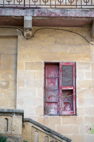 Eski bir bina yaşını gösterir. Kırmızı pencere pervazı soyulmuş ve cam kırılmış. Bina taştan yapılmış ve merdivenler pencereye çıkıyor..