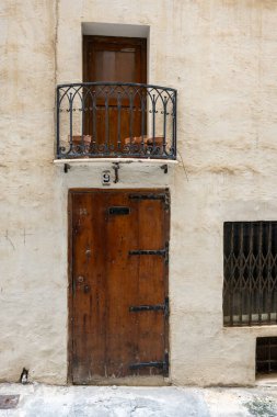 Ahşap kapılı eski bir binanın cephesi, saksıları ve parmaklıklı pencereleri olan küçük bir balkon. Mimari tarihi bir cazibeyi yansıtıyor..