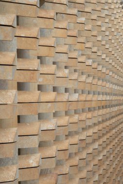 Açık kahverengi taş tuğlalardan oluşan dokulu bir duvar geometrik bir şablon yaratır. Tuğlalar derinlik ve gölge yaratacak şekilde istiflenmiş. Mimari tasarıma görsel ilgi katıyor..