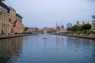 Birgu, Malta 'nın tarihi rıhtım manzarası. İnsanlar gezinti güvertesinde geziniyor, manzaranın ve limanın sakin sularının tadını çıkarıyorlar. Çeşme, sakin atmosfere katkı sağlar..