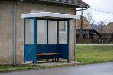 Kırsal bir yolda duran mavi bir otobüs durağı banliyö sakinlerine barınak sunuyor. Sessiz bir köy ortamında, doğa şartlarından korunan otobüsü beklemek için uygun bir yer..