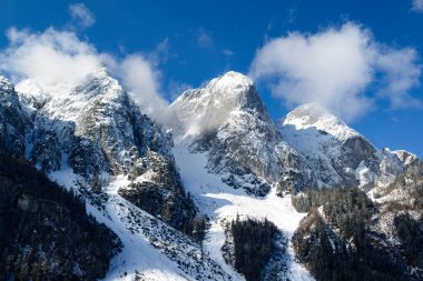 Görkemli, karla kaplı zirveler gökyüzüne uzanır, kayalık yüzleri beyaza boyanır. Zirvelerin etrafında dönen bulutlar dramatik bir alp manzarası yaratıyor..
