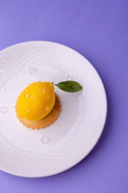 Aşırı gerçekçi limon şekilli bir tatlının yukarıdan çekimi: desenli yüzey, çiğ benzeri damlacıklar ve kıvrımlı beyaz bir tabakta yeşil yaprak..