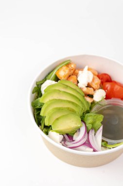 Geri dönüşümlü kağıt kasede canlı, katmanlı bir salata dilimlenmiş avokado, kızarmış tavuk, kiraz domatesleri, kırmızı soğan, roka ve yanında sos ile kremalı peynir içerir.. 