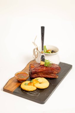 A slice of grilled meat with sauce, bread, and herbs on a stone board. Elegant plating in a restaurant setting against a white background.