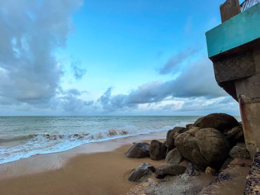 Ocean waves on sand near rocks. Cloudy sky and calm water create a dramatic, peaceful atmosphere.