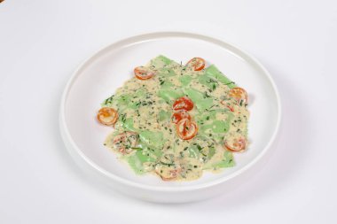 Bitki özlü makarnadan yapılan taze yeşil ravioli kremalı peynir sosu, yarım kiraz domatesleri ve beyaz bir tabakta doğranmış bitkilerle servis ediliyor. Minimalist sunum, stüdyo yemek fotoğrafçılığı.