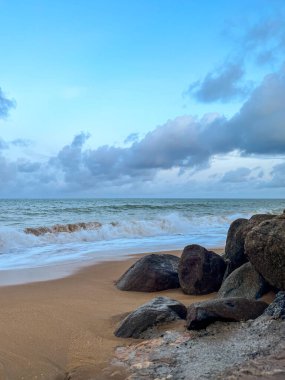 Ocean waves on sand near rocks. Cloudy sky and calm water create a dramatic, peaceful atmosphere.