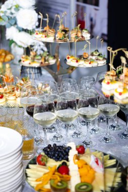 Kırmızı ve beyaz şarap bardakları, tepsilerde çeşitli kanepeler ve meyve ve peynir tabağı olan şenlikli bir büfe masasının yakın çekimi. Kutlama, düğün ya da kurumsal etkinlik için şık yemek organizasyonu..