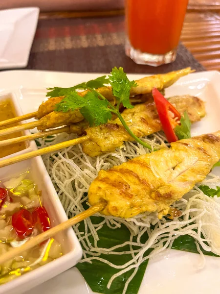 Grilled chicken skewers on bamboo sticks with sauce and herbs. Served on banana leaf and rice noodles.