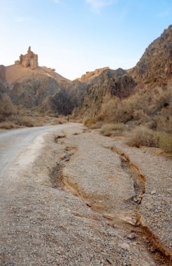 Bu fotoğraf engebeli kanyon duvarları ve kuru çalılar arasında uzanan dolambaçlı bir toprak yolu gösteriyor. Dramatik kanyon uçurumlarından geçen yol.