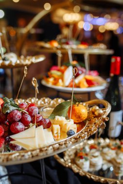 Taze üzümler, çilekler, portakallar, peynir dilimleri ve dekoratif yapraklarla dolu çok katlı zengin altın kaplama standlar altın sarısı detaylarla süslü zarif şişler, yumuşak sıcak bokeh ışıklandırması.