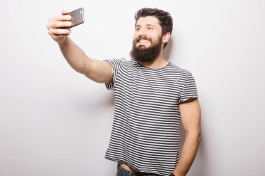 Beyaz izole arka plan üzerinde duran ve cep telefonu ile selfie çekerken sakallı gülümseyen adam.