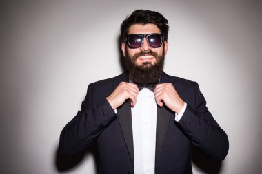 Güneş gözlüğü takan yakışıklı genç adamın, papyonunu ayarlayıp gri arka plana karşı dururken kameraya bakabak