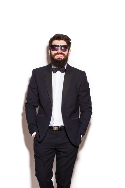Handsome young man wearing suit and glasses keeping hands in pockets and looking at camera while standing against white background — Stock Photo, Image