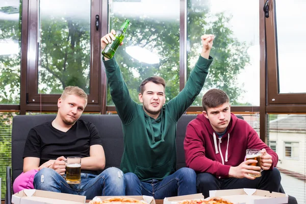 Τρεις ευτυχισμένος νεαροί άνδρες παρακολουθούν ποδόσφαιρο παιχνίδι και κρατώντας τα χέρια υψωμένα — Φωτογραφία Αρχείου