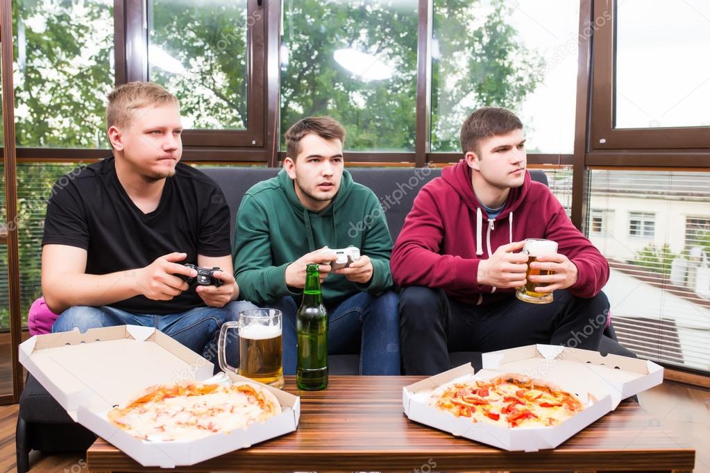 smiling male friends playing video games, drink beer and have fun at home