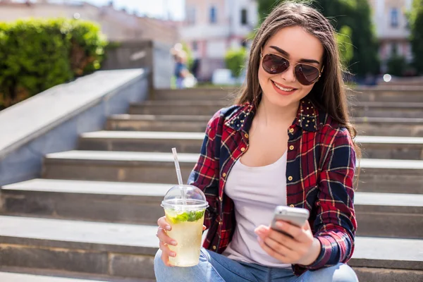 Mujer con una taza fresca sentada en las escaleras y usando su smartphone para comunicarse . — Foto de Stock