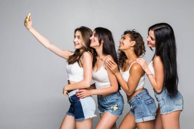 Mutlu gülümseyen dört genç kız beyaz arka planda izole bir şekilde telefonla selfie çekiyor..