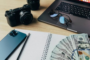 Kamera, defter, gözlük, dizüstü bilgisayar, akıllı telefon ve dolar banknotları olan ofis masası..