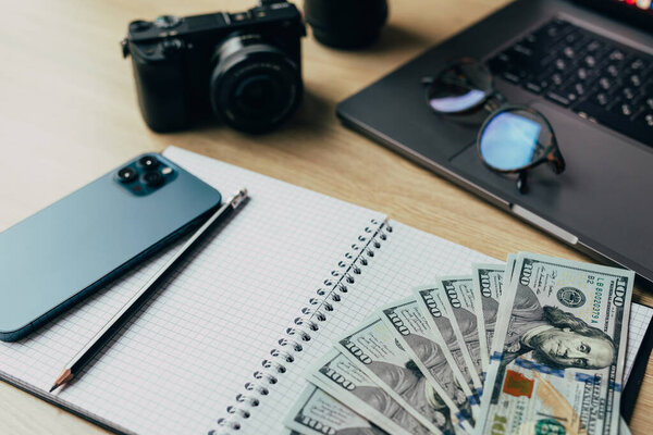 Office desk workplace with camera, notebook, glasses, laptop, smartphone and dollars banknotes on wooden table background.