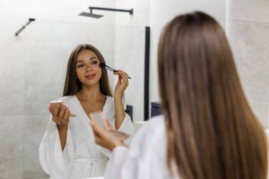 Gülümseyen genç bir kadın yüzüne makyaj yapıyor beyaz bornozlu kozmetik fırçasıyla sabah hazırlıkları yapıyor. Güzellik ve Tedavi