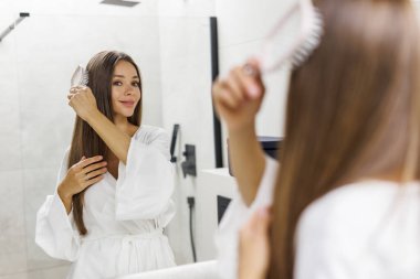 Banyoda maske taktıktan sonra saçını tarayan genç bir kadın.