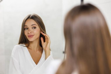 Cilt Kozmetik. Genç bir kadın banyo yaptıktan sonra nemlendirici krem sürüyor. Küvette oturuyor, modern banyoda beyaz bornoz giyiyor. Kadın güzellik rutini