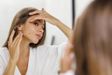 Banyoda duran bir kadın lavabo musluğuyla aynaya bakarak yanaklarına dokunuyor. Özbakım, bakım, cilt bakımı, sükunet, zarafet, minimalizm, rahatlama.