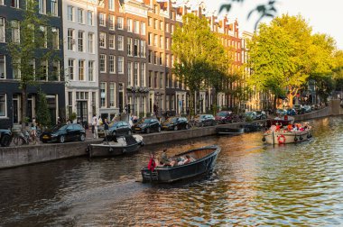 Amsterdam, Hollanda - 14 Temmuz 2025: Tarihi binalar ve botlar sıcak bir günbatımı gökyüzü altında sakin kanal sularına yansıdı.