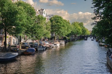 Amsterdam, Hollanda - 14 Temmuz 2025: Tarihi binalar ve botlar sıcak bir günbatımı gökyüzü altında sakin kanal sularına yansıdı.