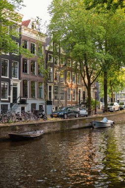Amsterdam, Hollanda - 14 Temmuz 2025: Tarihi binalar ve botlar sıcak bir günbatımı gökyüzü altında sakin kanal sularına yansıdı.