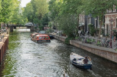 Amsterdam, Hollanda - 14 Temmuz 2025: Tarihi binalar ve botlar sıcak bir günbatımı gökyüzü altında sakin kanal sularına yansıdı.