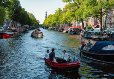 Amsterdam, Hollanda - 14 Temmuz 2025: Tarihi binalar ve botlar sıcak bir günbatımı gökyüzü altında sakin kanal sularına yansıdı.