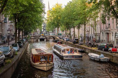 Amsterdam, Hollanda - 14 Temmuz 2025: Tarihi binalar ve botlar sıcak bir günbatımı gökyüzü altında sakin kanal sularına yansıdı.