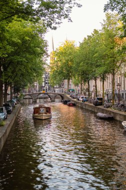 Amsterdam, Hollanda - 14 Temmuz 2025: Tarihi binalar ve botlar sıcak bir günbatımı gökyüzü altında sakin kanal sularına yansıdı.