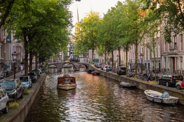 Amsterdam, Hollanda - 14 Temmuz 2025: Tarihi binalar ve botlar sıcak bir günbatımı gökyüzü altında sakin kanal sularına yansıdı.