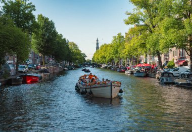 Amsterdam, Hollanda - 14 Temmuz 2025: Tarihi binalar ve botlar sıcak bir günbatımı gökyüzü altında sakin kanal sularına yansıdı.